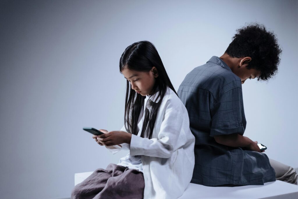 A imagem com uma paleta de cores cinza, azul e branco, mostra dois adolescentes de costas um para o outro, com olhares distantes sem interagir, mas conectados com seu celulares.
